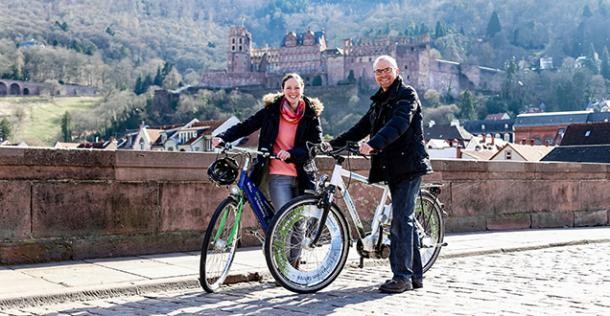 Heidelberg hat die meisten Fahrrad-Fahrer (Foto: Stadt Heidelberg/ Pellner) Eine Frau und ein Mann stehen mit zwei Fahrrädern auf der Alten Brücke. Im Hintergrund ist das Schloss und die Sonne scheint. (Foto: Stadt heidelberg/ Pellner)