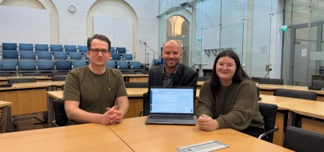 Zu sehen sind 3 Personen die im Großen Sitzungsaal sitzen und einen Laptop präsentieren.