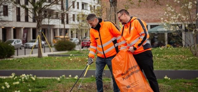 Zwei Männer sammeln mit Greifzangen Müll vom Boden auf. Sie tragen orangene Warnjacken.