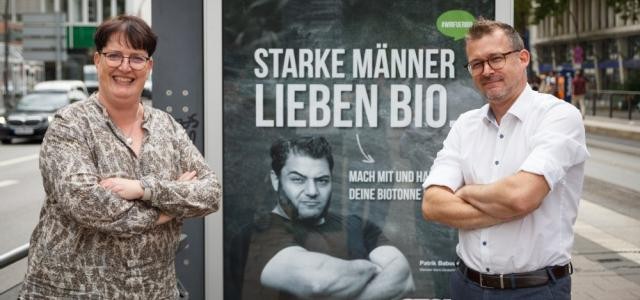 Sylvia Hafner und Umweltbürgermeister Raoul Schmidt-Lamontain stehen vor der City-Light-Säule mit dem Aufdruck: "Starke Männer Lieben Bio", begleitet wird der Aufdruck von dem Bild eines Arme verschränkenden, stark aussehenden Mannes.