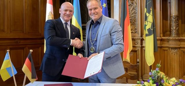 Zwei Herren, in einem schönen holzvertäfelten Saal, geben sich die Hand und halten eine Urkunde in die Kamera. Hinter ihnen  die Flaggen der Stadt Odessa, der Ukraine, der Europäischen Union, der Bundesrepublik Deutschlands und des Landes Baden-Württemberg.