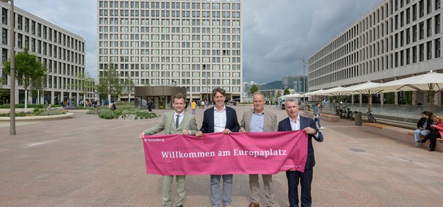 Vier Männer in Anzug halten ein heidelbeerfarbenes Banner mit der Aufschrift "Willkommen am Europaplatz"
