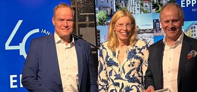 Eine Frau und zwei Männer posieren mit einer gerahmten Urkunde.