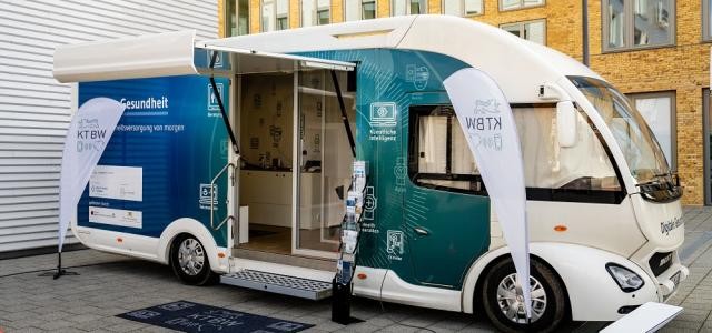 Ein futuristisch aussehender Bus steht vor einem Gebäude. Man kann in den Bus hineingehen.