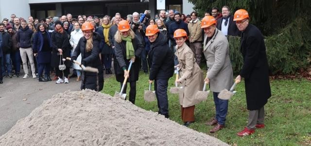 Sechs Menschen halten Spaten in den Händen, mit denen sie Erde aufgenommen haben. Sie schauen in die Kamera, hinter ihnen steht Publikum.