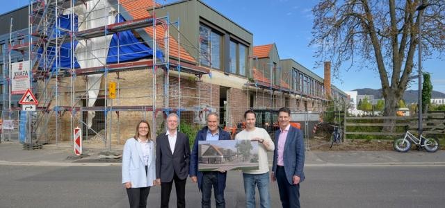 Vier Männer und eine Frau stehen vor einem Gebäude und lächeln in die Kamera. Zwei der Männer halten ein Schild in den Händen.