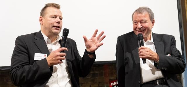 Zwei Männer sitzen auf einer Bühne und halten jeweils ein Mikrofon in der Hand. Sie unterhalten sich, einer von ihnen lacht.