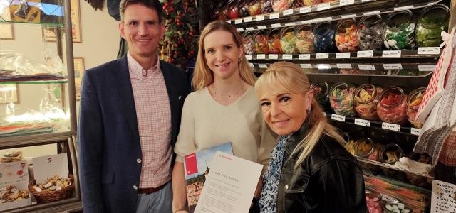 Ein Mann und zwei Frauen lächeln in die Kamera, die Frau in der Mitte hält ein Schriftstück in den Händen.