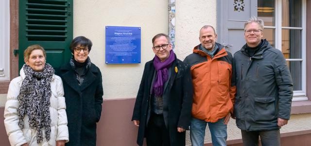 Fünf Personen posieren vor einer blauen Gedenktafel an einer Hauswand.