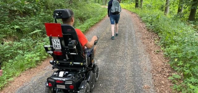 Mann fährt mit E-Rolli auf einem Waldweg mit einer Begleitung, die vorausgeht.
