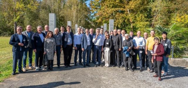 Eine gruppe von Menschen steht auf einem Schotterweg vor dem von Bäumen umsäumten Deportiertenfriedhof.