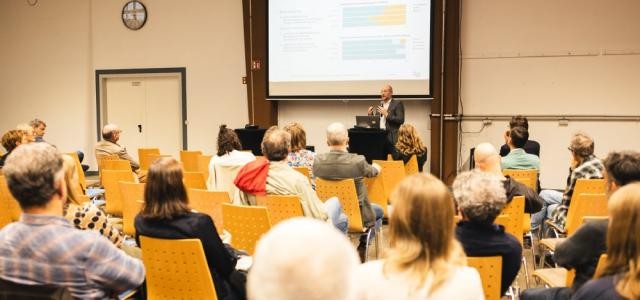Eine Gruppe von Menschen sitzt in Reihen und betrachtet vor sich eine Präsentation die von einem Mann gegeben wird