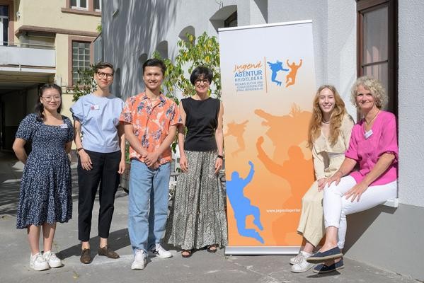 Die Kompetenzagentur der Jugendagentur Heidelberg unterstützt benachteiligte Jugendliche (v.l.): Jenny Li (Team Kompetenzagentur), Caroline Smout (Amt für Chancengleichheit der Stadt Heidelberg), Projektteilnehmer Nemat, Bürgermeisterin Stefanie Jansen, Projektteilnehmerin Natalie und Bettina Kolbe (Team Kompetenzagentur) (Foto: Rothe). Sechs Menschen stehen vor einem Gebäude neben einem orangefarbenen Roll-up in der Sonne