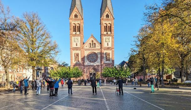 Angeleitete Bewegungsübungen im Freien zusammen mit anderen Menschen aus dem Stadtteil: das konnten ältere Menschen beim Aktionstag zum zehnten Jubiläum der Bewegungstreffs auf dem Wilhelmsplatz in der Weststadt testen. (Foto: Stadt Heidelberg) Ältere Menschen machen Dehnübungen vor einer Kirche.