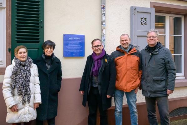 Stefanie Jansen, Bürgermeisterin für Soziales, Bildung, Familie und Chancengleichheit (2. von links), mit Hauseigentümerin Ingrid Longerich (von links), Helmut Metzner, Geschäftsführender Vorstand des Bundesstiftung Magnus Hirschfeld, Ideengeber Steffen Schmid und Stefan Kaumkötter vom Kulturamt der Stadt Heidelberg vor der neuen Gedenkplakette für Magnus Hirschfeld in der Sandgasse. Fünf Personen posieren vor einer blauen Gedenktafel an einer Hauswand.