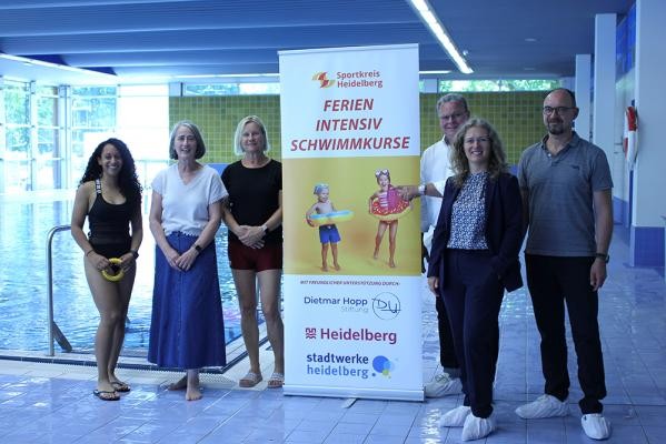 Freuen sich über die Kooperation im Rahmen der kostenlosen Ferienschwimmkurse (v.l.): Daniela Pfeuti, Projektleiterin Schwimmkurse beim Sportkreis Heidelberg, Heike Bauer, Leiterin der Dietmar Hopp Stiftung, Annette Kietzmann, Koordinatorin der Schwimmlehrkräfte beim ISSW, Gert Bartmann, Leiter des Amts für Sport und Gesundheitsförderung, Maike Carstens, Geschäftsführerin der Stadtwerke Heidelberg Bäder, und Ralph Fülop, Geschäftsführer beim Sportkreis Heidelberg. (Foto: Stadt Heidelberg) Sechs Personen stehen in einem Schwimmbad um einen Aufsteller mit der Aufschrift "Ferien Intensiv Schwimmkurse".