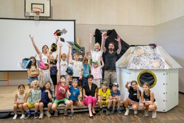 Galaktischen Ferienspaß und eine große Talentshow konnte Bürgermeisterin Stefanie Jansen (vordere Reihe Mitte sitzend) auf ihrer Sommertour mit den Kindern bei der Ferien-Spielaktion im Haus der Jugend erleben. (Foto: Philipp Rothe) Bürgermeisterin Stefanie Jansen sitzt mit den Kindern und Mitarbeitenden der Ferien-Spielaktion in der Turnhalle im Haus der Jugend.