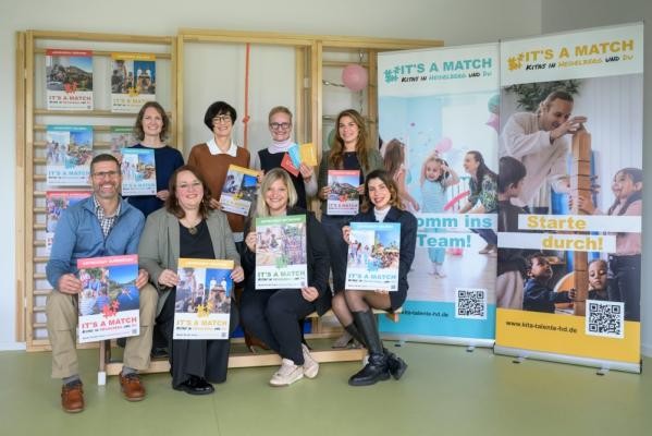 Werben um engagierte Fachkräfte für die mehr als 130 Heidelberger Kitas: Bürgermeisterin Stefanie Jansen (hinten, 2. von links) und Jugendamtsleiterin Myriam Lasso (hinten, 3. von links) mit Kita-Fachkräften und Vertreterinnen des Kinder- und Jugendamts sowie der Agentur Commha Consulting. (Foto: Philipp Rothe) Bürgermeisterin Stefanie Jansen steht mit Myriam Lasso und 6 weiteren Personen vor einer Holzkletterwand für Kinder und neben zwei Werbeaufstellern für It´s a match.