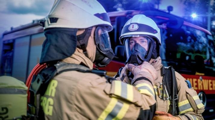 In der zehnten Staffel der Doku-Reihe „Feuer & Flamme“ können die Zuschauerinnen und Zuschauer hautnah die Einsätze der Feuerwehr Heidelberg verfolgen. (Foto: SWR/Sebastian Drolshagen) Zwei Feuerwehrpersonen stehen vor einem Löschfahrzeug. Einer legt dem anderen eine Gasmaske an.