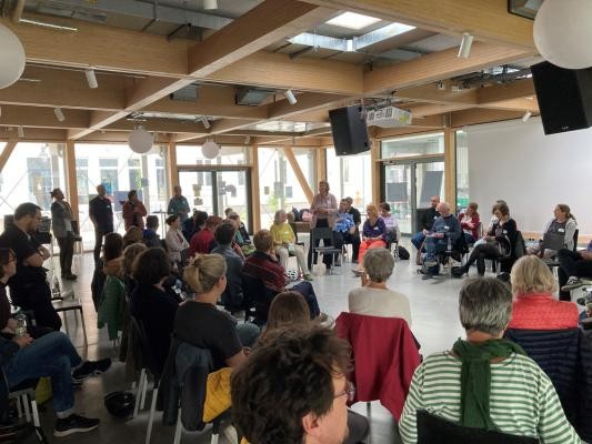 Gemeinschaftliche Wohnprojekte werden immer beliebter: Das Vernetzungstreffen in der Aula des Collegium Academicum im Mai 2025 war gut besucht. (Foto: Stadt Heidelberg) Eine Gruppe von Menschen sitzt kreisförmig in einem gut durchlichteten Raum zusammen und unterhält sich.