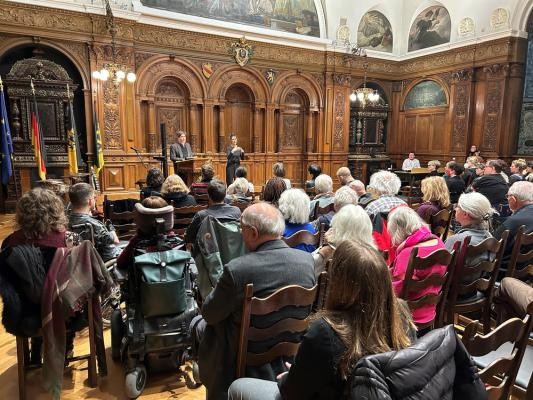 „Das Erinnern und Gedenken an die Verbrechen des Nationalsozialismus ist eine besondere Verant-wortung. Gerade in der aktuellen politischen Lage, in der Hass und Hetze zunehmen, ist es wichtig, dass wir mit offenen Augen durch unsere Stadt gehen“, mahnte Erste Bürgermeister Jürgen Odszuck in seiner Rede bei der Gedenkveranstaltung am 27. Januar 2026. Viele Personen sitzen auf Stühlen, zwei Personen stehen