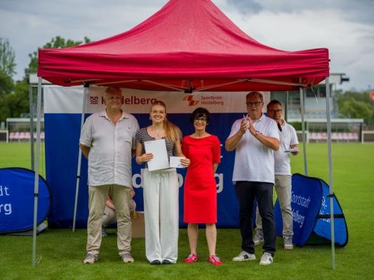 Bei der Wahl zu Heidelbergs Jugendsportlerin des Jahres wurde Irmela Zeh vom SV Nikar Heidelberg Zweite (auf dem Bild mit von links Dr. Peter Schlör, Vorsitzender des Sportkreises Heidelberg, Bürgermeisterin Stefanie Jansen und Gert Bartmann, Leiter des Amtes für Sport und Gesundheitsförderung). Die Erstplatzierte bei der Jugendsportlerinnenwahl – Basketballerin Leonie Pfanzelt vom Olympiastützpunkt – konnte leider nicht an der Auszeichnung teilnehmen. (Foto: Tobias Dittmer) Jugendsportlerin Irmela Zeh wird vor einem Zelt geehrt.