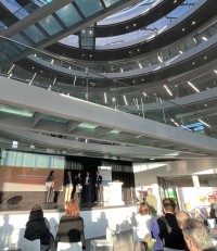 Cellzome lud ins beeindruckende Advanced Training Center am EMBL ein. (Foto: Stadt Heidelberg) Mehrere Menschen stehen auf einer Bühne. Über ihnen führen Glasstege in die Höhe. Vor ihnen sitzt Publikum.