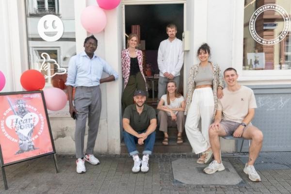 Präsentieren im neuen Konzeptladen HeartWork ihre Produkte (von links): Madiagne Diop, Lena Grunder, Oliver Matlock, Simon Fritz, Alexandra Quandt, Sofia Leser und Moritz Stock. (Foto: Larionova) Mehrere Menschen sitzen, stehen und hocken vor einem Laden, der mit pastellfarbeenen Luftballons geschmückt ist.