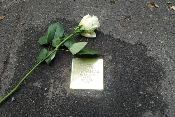 Ein messingfarbener Stolperstein in Pflastersteingröße ist n den Boden eingelassen, flankiert von einer weißen Rose
