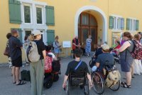 Eine Gruppe von Menschen, stehend oder im Rollstuhl, vor einem gelben Gebäude.